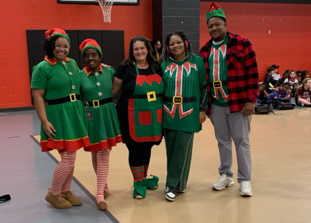 Five people in elf costumes stand in a gymnasium with a basketball hoop visible in the background.