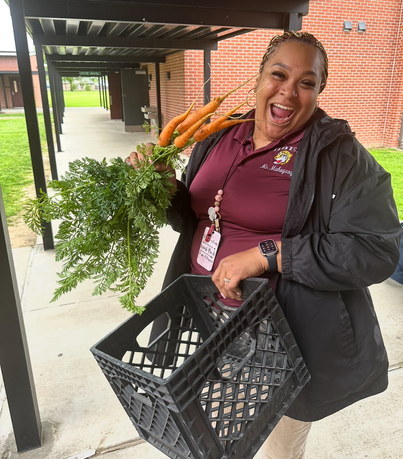A joyful woman in the foreground holds a bunch of carrots and a milk crate, with a brick building and outdoor walkway in the background.
