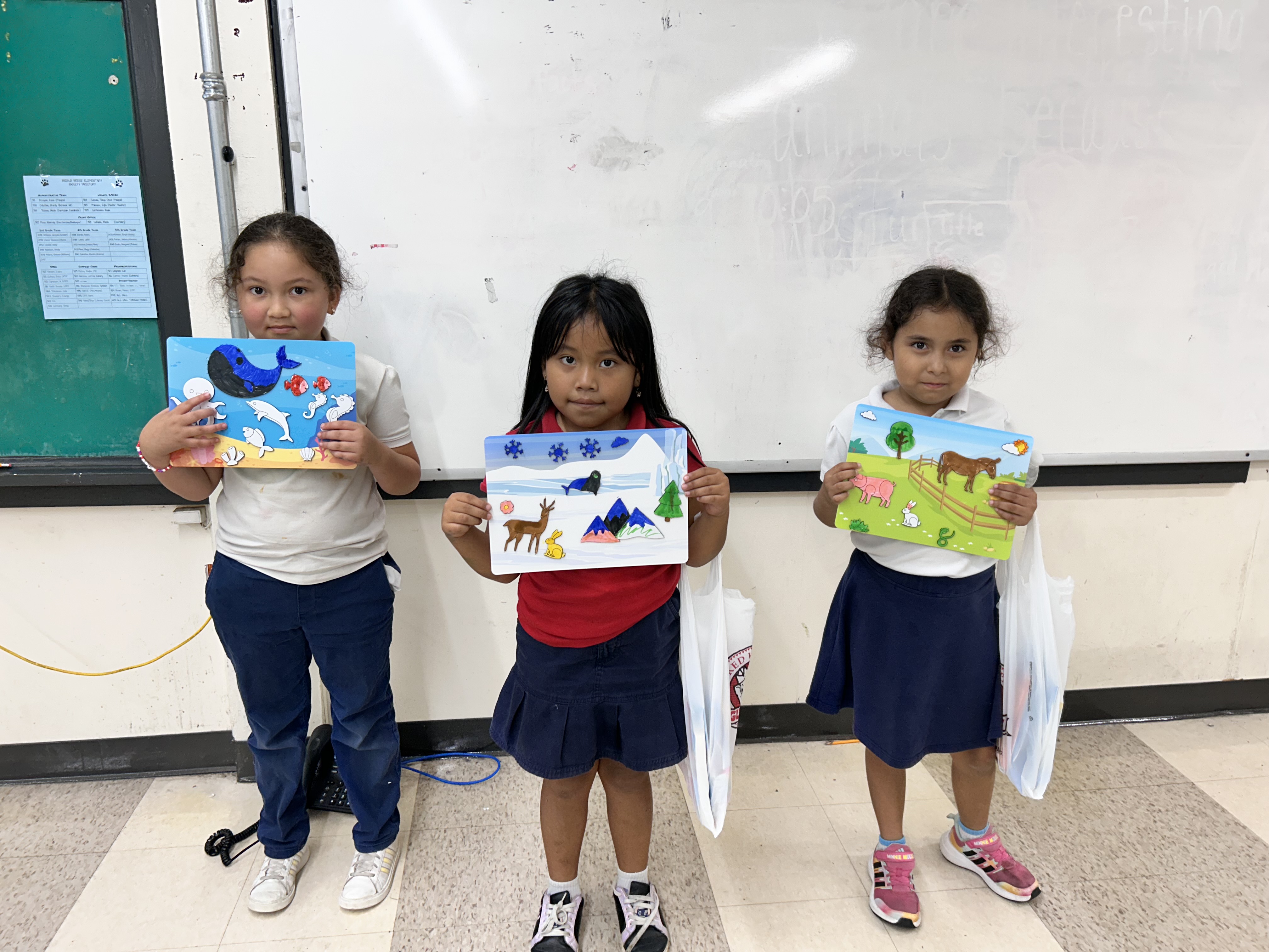 Three young girls stand in a classroom holding up their artwork, with a whiteboard and a bulletin board visible in the background.