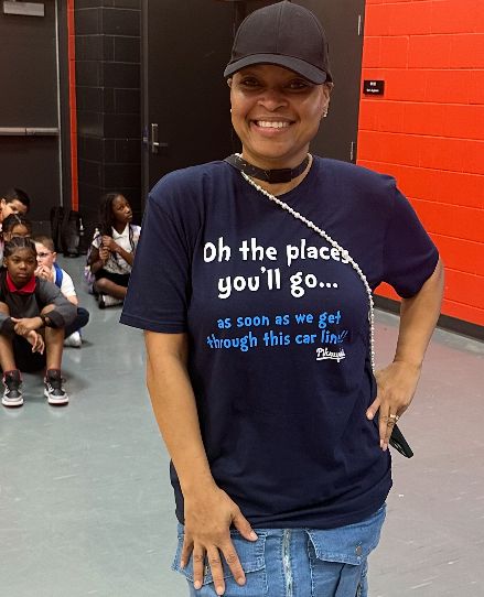 A woman in a dark blue t-shirt with white text smiles at the camera in the foreground, while a group of children sit on the floor in the background.