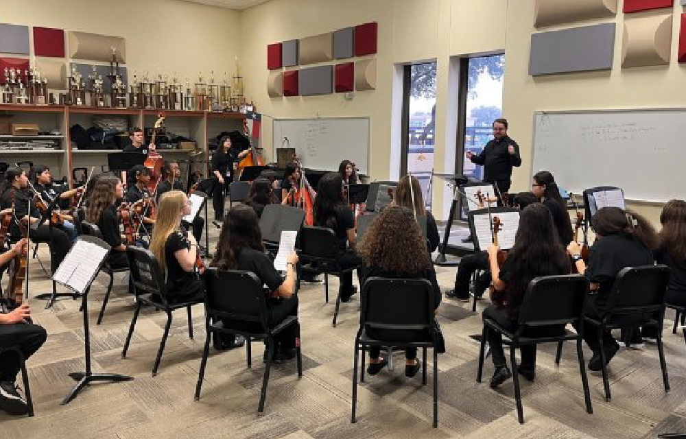 A performance or rehearsal is taking place in a classroom-like setting, with a group of musicians and performers seated and standing, and various musical instruments and equipment visible in the background.