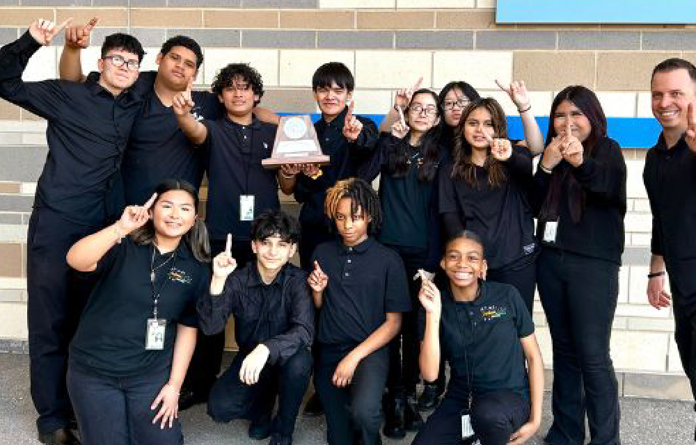 A group of people, all wearing black uniforms, are gathered together and making various hand gestures, with a brick building visible in the background.