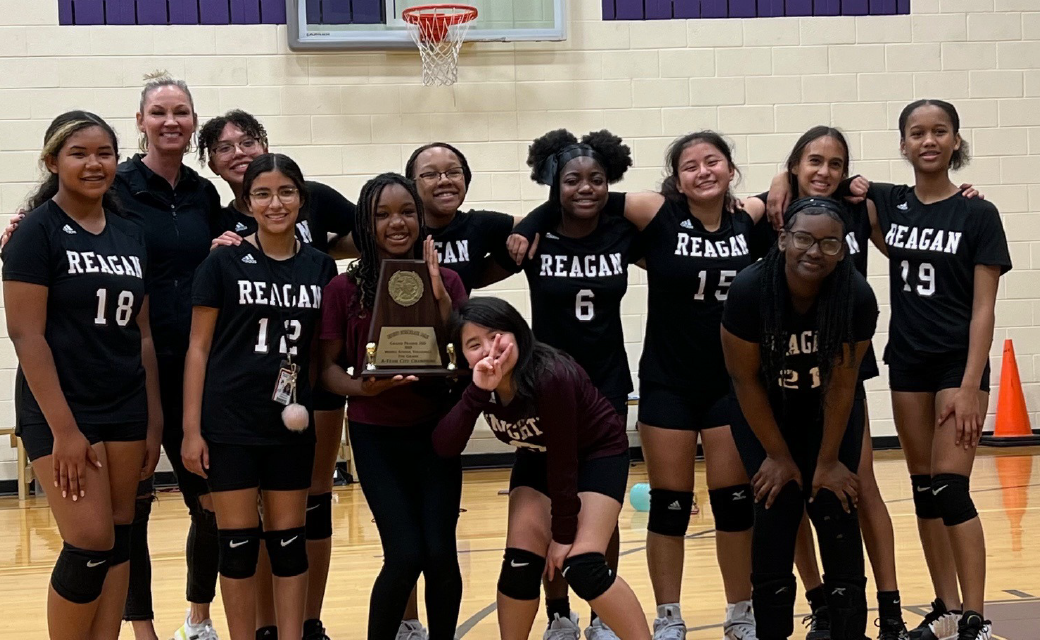 A group of young female basketball players in black uniforms with "Reagan" printed on them, posing together and holding a trophy in a gymnasium setting with a basketball hoop visible in the background.