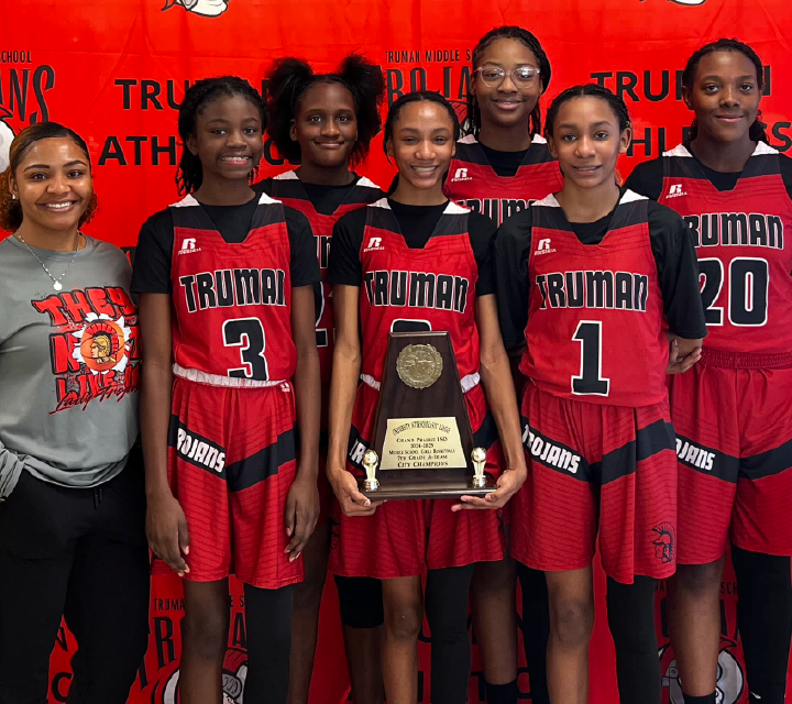 A group of young female basketball players in red uniforms holding a trophy, standing in front of a red background with the word "Truman" prominently displayed.