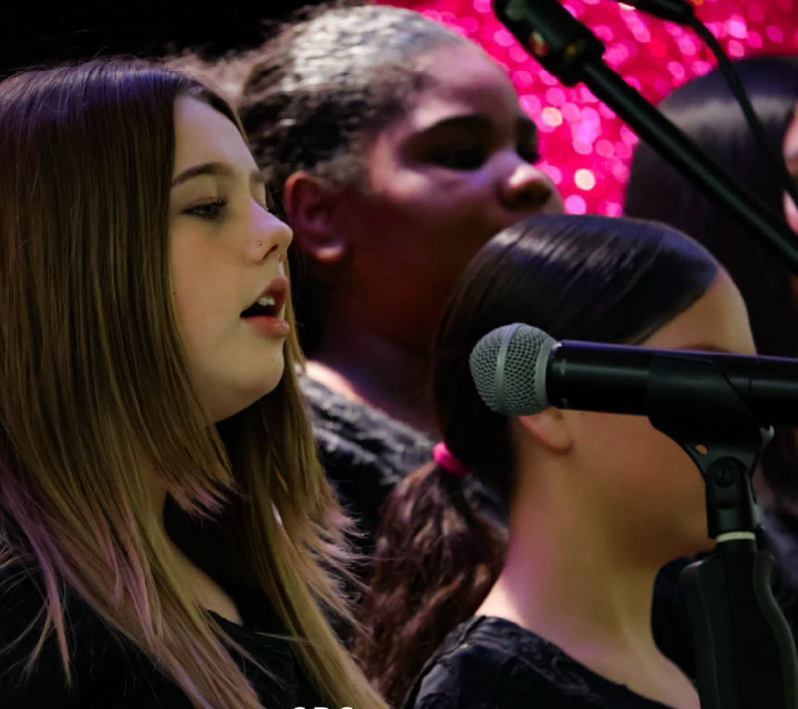 A woman with long, dark hair is singing into a microphone, while a man with a beard is playing a guitar in the background, with colorful lights illuminating the scene.