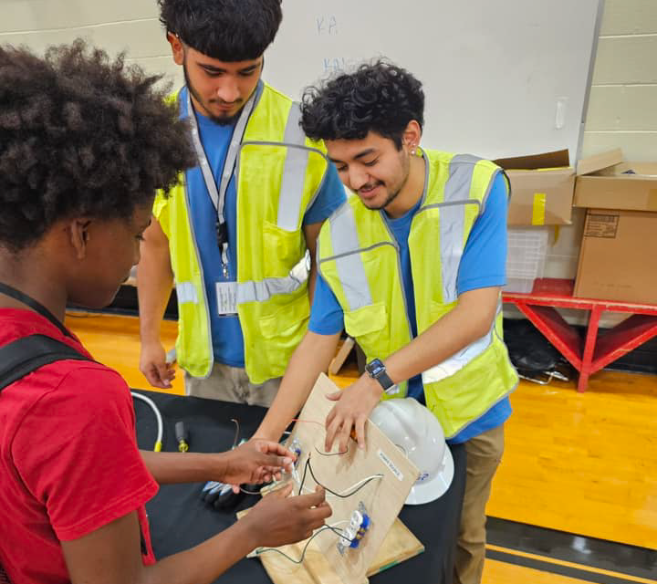 The image shows three individuals wearing high-visibility vests, engaged in what appears to be a hands-on activity or demonstration. The background suggests a warehouse or industrial setting, with various equipment and storage containers visible.