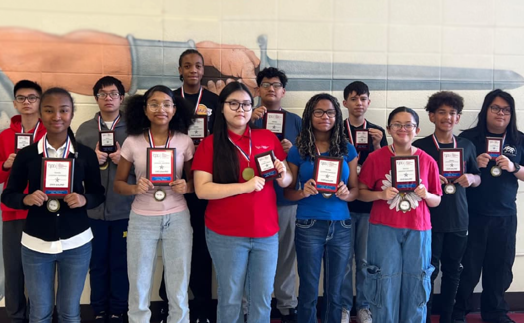A group of young people, mostly women, are standing together and holding up what appear to be awards or certificates in a setting with a large building or structure in the background.
