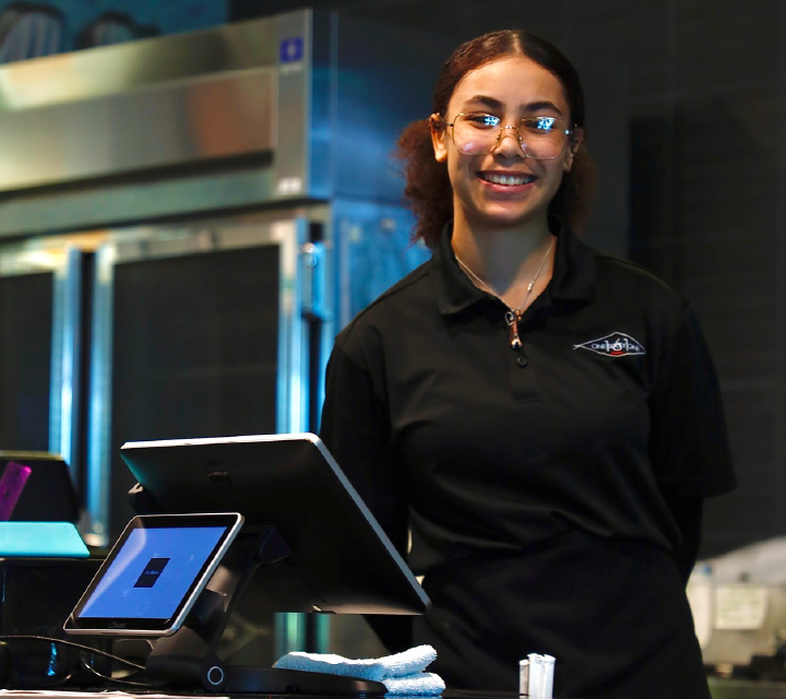 A smiling young woman in a black shirt stands in front of a computer workstation with various electronic devices visible in the background.