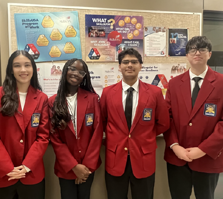 A group of four students wearing red blazers stand in front of a bulletin board displaying various educational and career-related information.