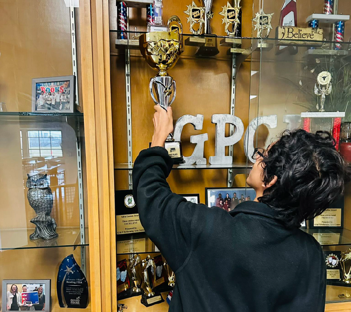 The image shows a person in a black jacket reaching up to touch the letters "GPO" displayed on a shelf filled with various trophies and memorabilia.