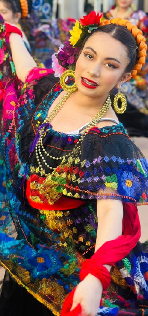 A vibrant and colorful woman in traditional Mexican attire, adorned with flowers and jewelry, stands against a backdrop of equally vibrant and patterned fabrics.