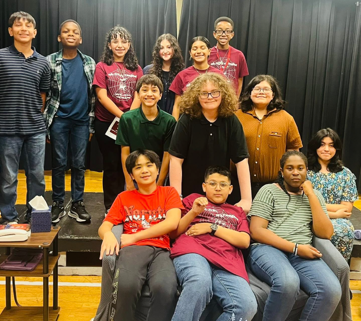 A group of diverse individuals, both adults and children, are gathered together in what appears to be a school or community setting, with a stage or performance area visible in the background.