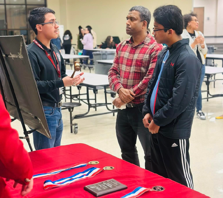 Three men are engaged in conversation in what appears to be a classroom or educational setting, with a red table in the foreground displaying various items.