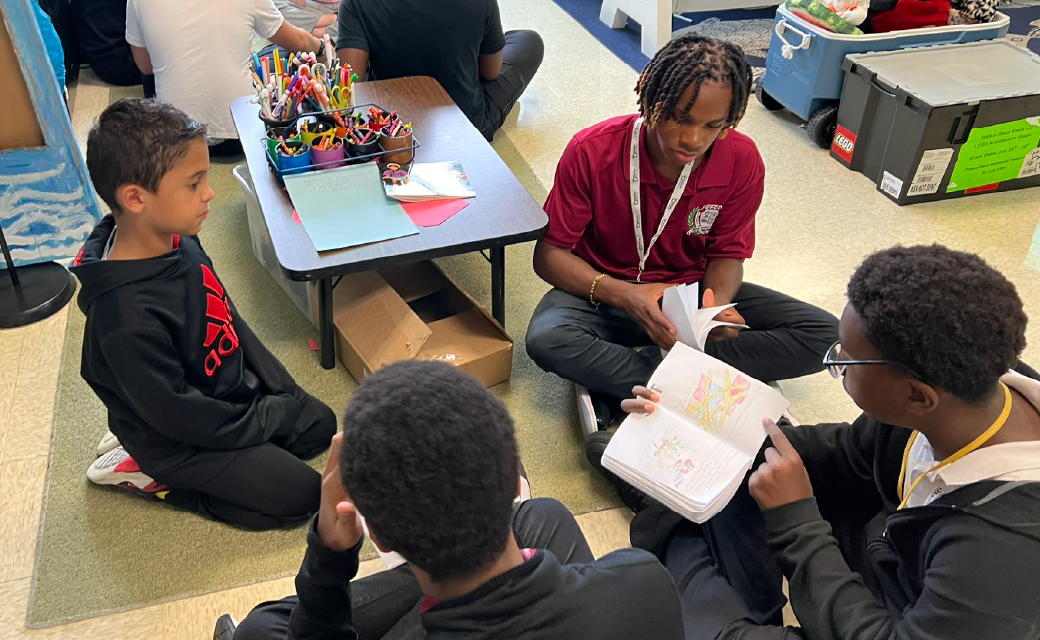 A group of students sitting around a table, engaged in a collaborative learning activity, with various educational materials and supplies visible in the background.
