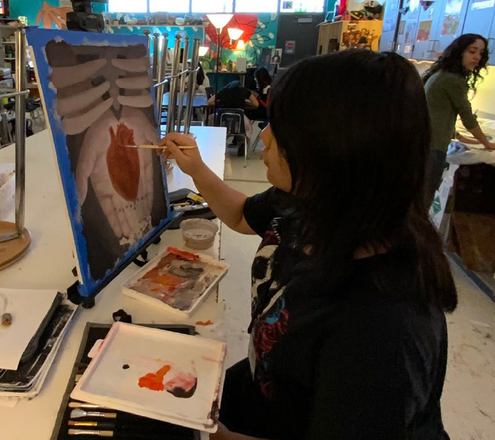 A person in a dark jacket is painting on an easel in a cluttered classroom setting, with various art supplies and decorations visible in the background.