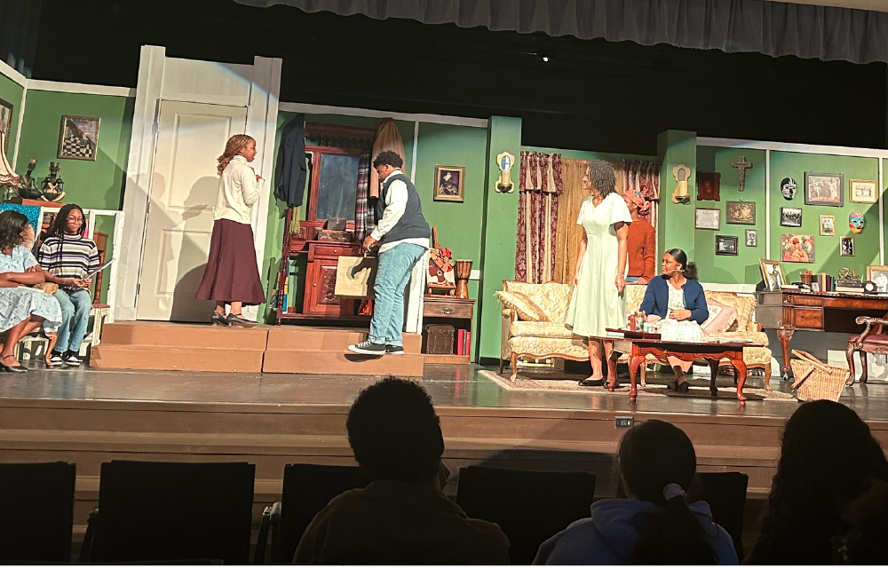 The image depicts a stage with several actors performing a theatrical production, with the audience visible in the foreground.
