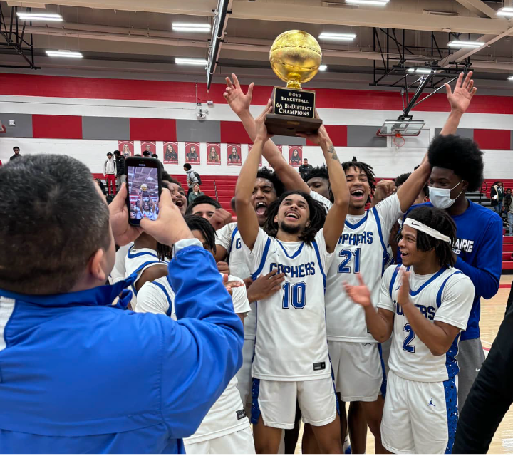 A group of basketball players in blue and white uniforms are celebrating and holding up a trophy in a gymnasium setting with red and white walls.