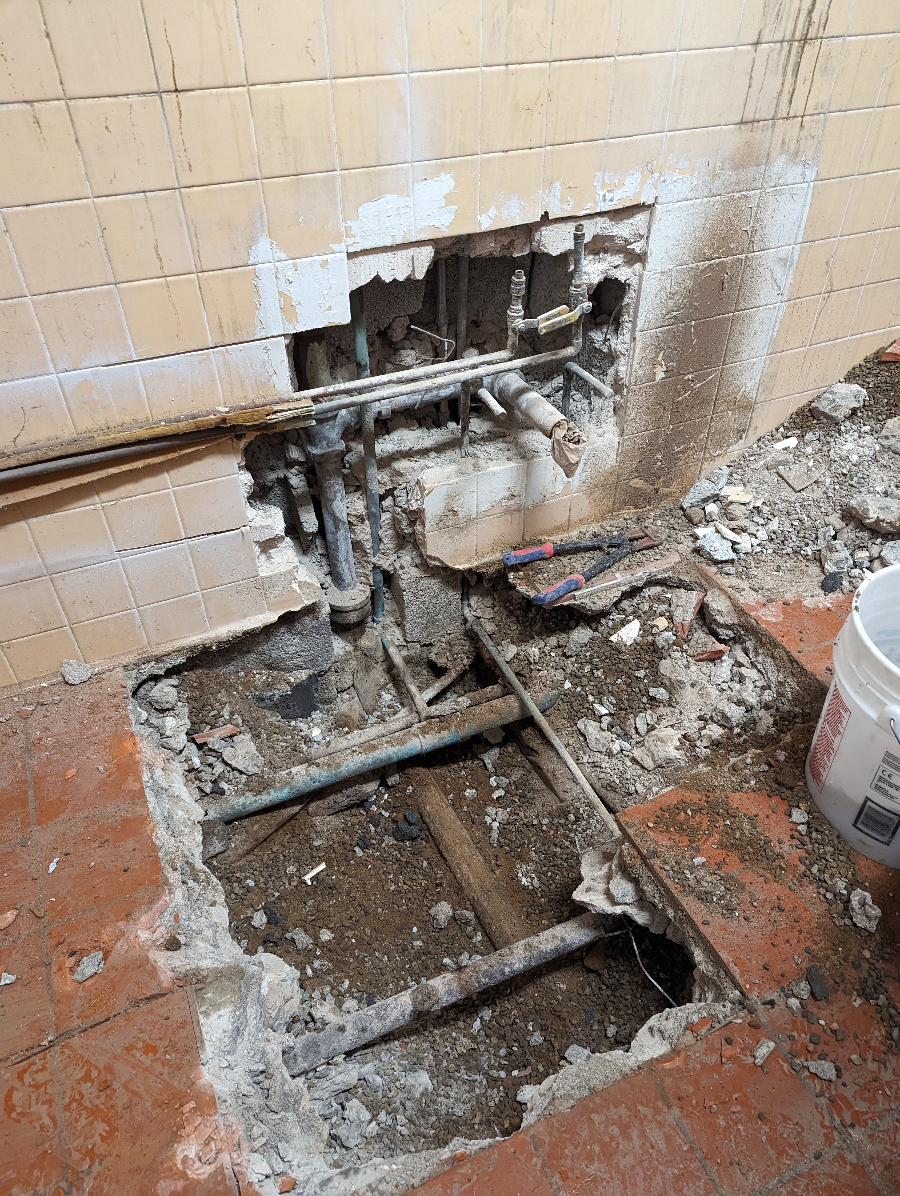 Exposed plumbing and debris fill a hole in the floor in front of a tiled wall with a section of plaster removed to reveal more pipes.