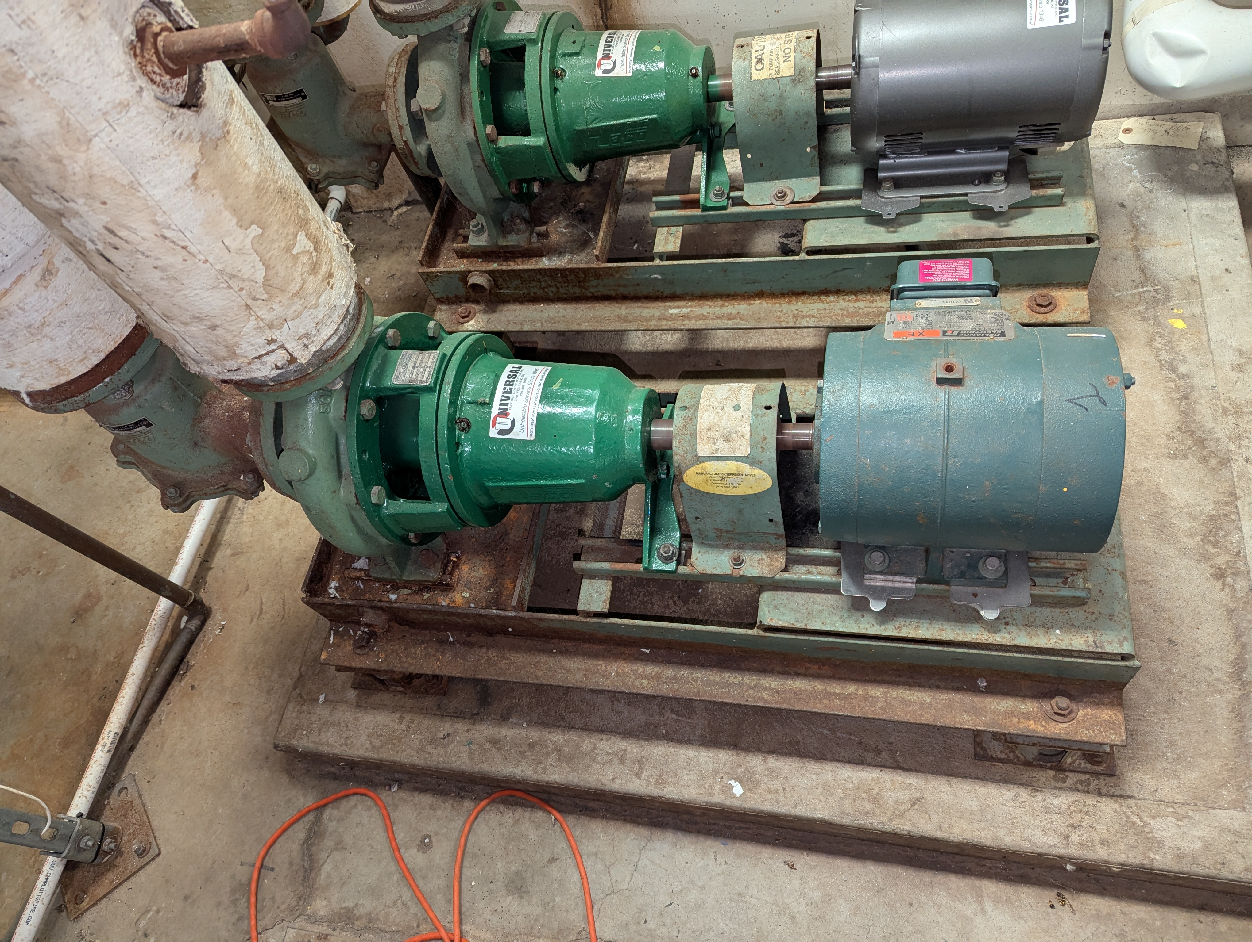 Two industrial pumps with green casings and electric motors are mounted on metal bases in the foreground, with pipes extending from the left and a concrete floor and wall in the background.