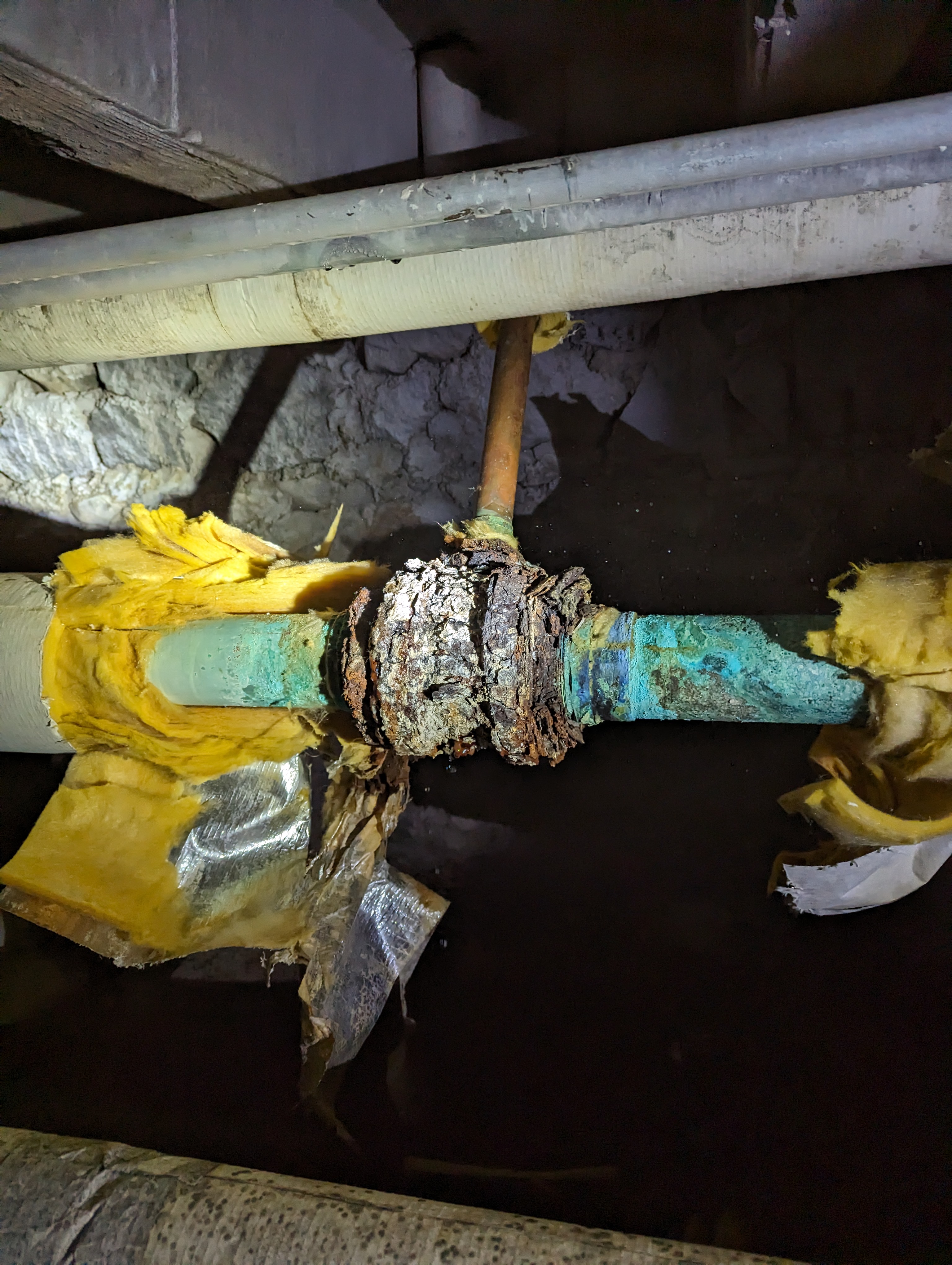 Corroded copper pipes with insulation are visible in the foreground, set against a dark, rough background of what appears to be a crawl space.