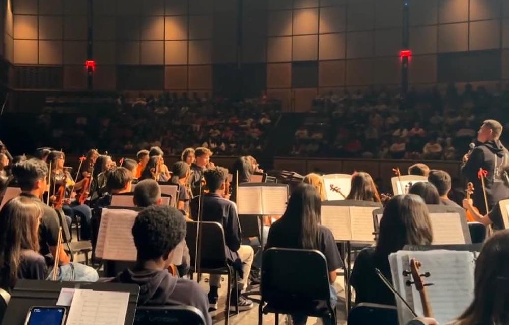 A large audience seated in a dimly lit auditorium, with a stage and orchestra visible in the background.