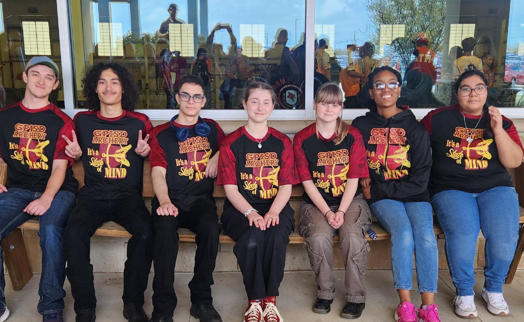 The image shows a group of young people, likely a team or club, wearing matching t-shirts and posing together in front of a mural or artwork on the wall behind them.