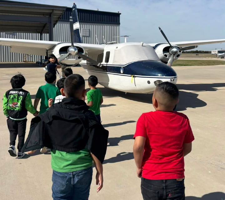 A group of children in colorful shirts are gathered around a large propeller-driven aircraft, likely an airplane, parked on a paved surface with buildings in the background.