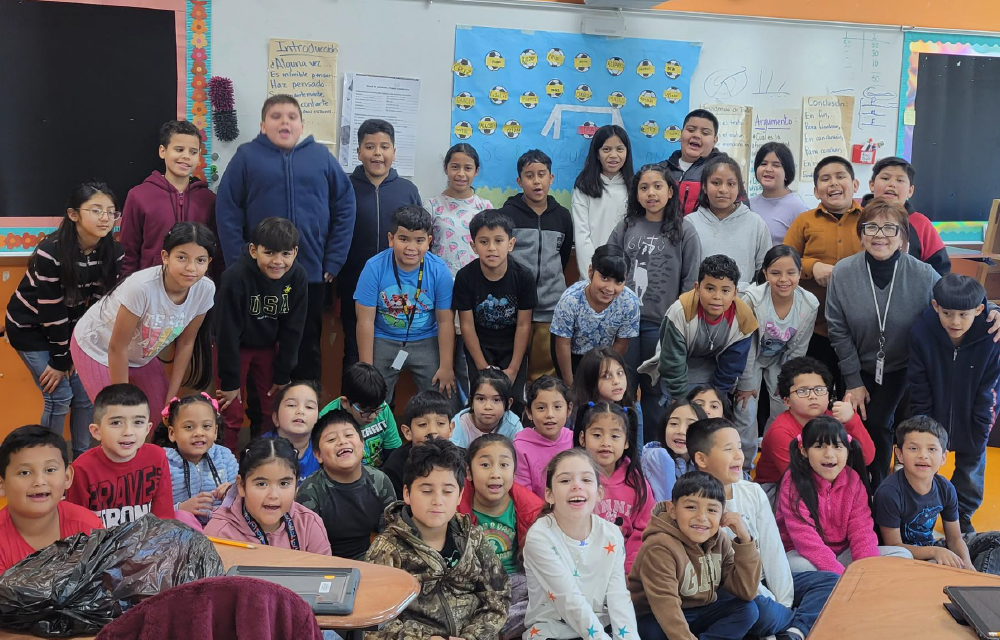 A large group of children and adults, likely students and teachers, gathered in a colorful classroom setting with various educational materials and displays visible in the background.