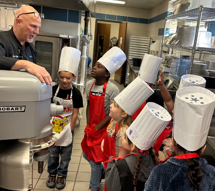 The image shows a group of young children, dressed in chef's uniforms, standing in a commercial kitchen and interacting with an adult chef.