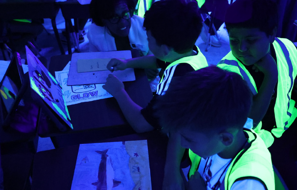The image depicts a group of people in a dark, neon-lit environment, with various glowing and illuminated objects and signs visible in the background.