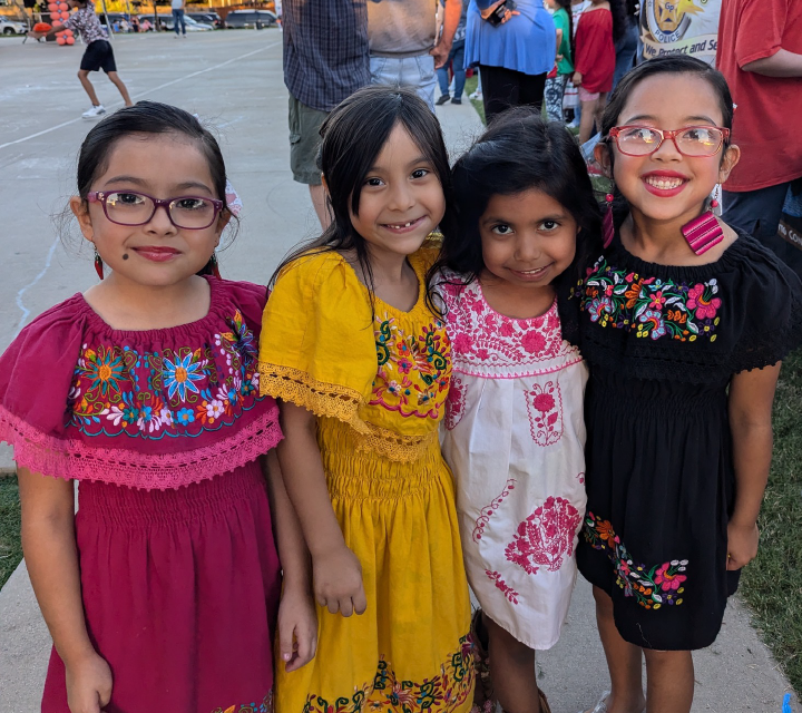 Four young girls, dressed in colorful traditional Mexican dresses, are standing together and smiling at the camera. In the background, there appears to be a street scene with various people and buildings.