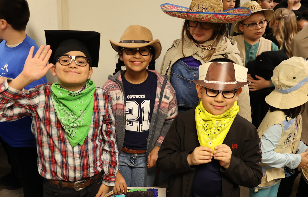 A group of people wearing various costumes and accessories, including hats, sunglasses, and bandanas, posing together in what appears to be a classroom or similar indoor setting.