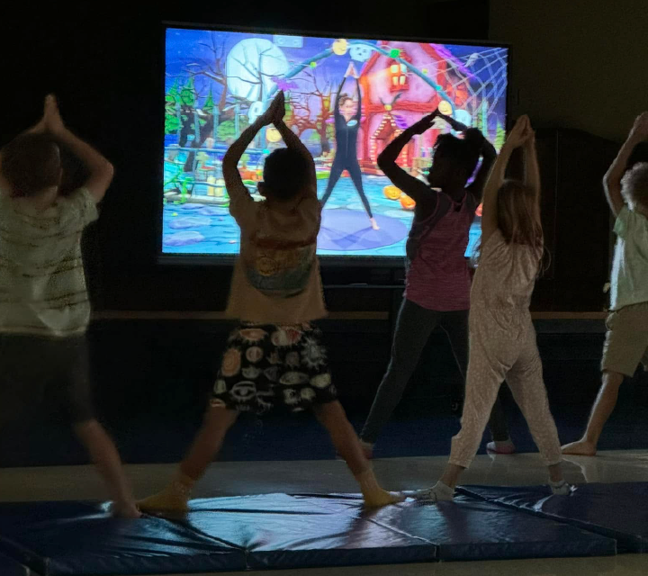 The image shows several people performing dance moves in front of a large, colorful, and animated projection screen displaying a whimsical, fantastical scene.