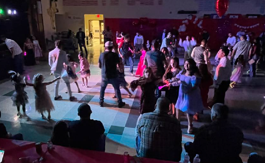 A crowded dance floor with people dressed in formal attire, some dancing and others standing around the edges of the room.