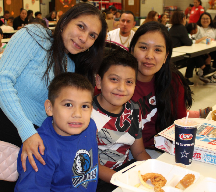 A group of smiling people, including two women and two children, are seated at a table in what appears to be a cafeteria or dining area.