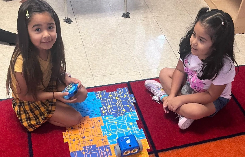 Two young girls, appearing to be elementary school-aged, are sitting on a colorful patterned rug in what seems to be a classroom or educational setting. They are engaged in some kind of activity or play, with one girl holding a handheld device.