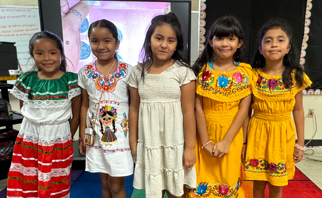 A group of young girls wearing colorful, traditional Mexican-inspired dresses stand together in what appears to be a classroom or educational setting, with a whiteboard and other classroom items visible in the background.