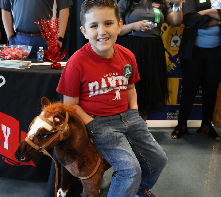 A young boy in a red t-shirt is sitting on a stuffed horse toy, surrounded by various other toys and items in what appears to be a gaming or entertainment setting.