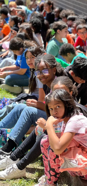 A large crowd of people, mostly young, sitting on the ground in a park-like setting, engaged in various activities and interactions.