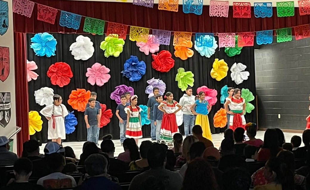 A colorful stage with vibrant paper flowers and decorations, featuring a group of performers in traditional Mexican attire, entertaining an audience seated in the foreground.