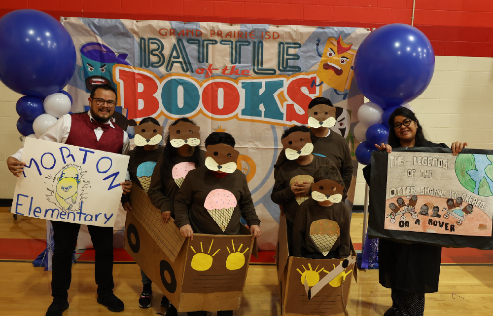 The image shows a group of people, likely students, posing in front of a large banner promoting a "Battle of the Books" event. The people are wearing costumes or holding signs related to books, and the background includes balloons and other decorations, suggesting this is a school or library event.