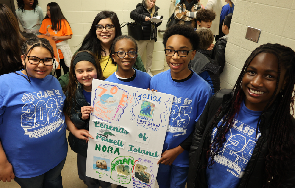 A group of smiling young people wearing blue shirts with graphics, holding a colorful poster, and standing in what appears to be a school or community setting.