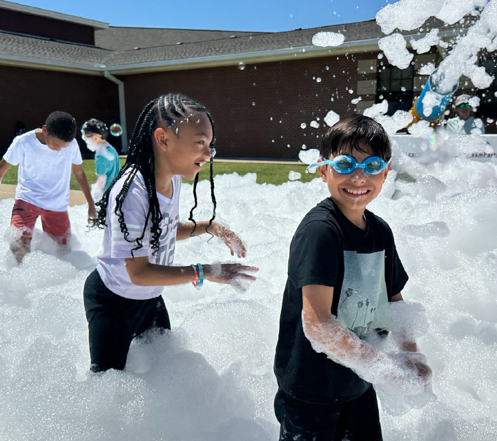 Two young children, a boy and a girl, are playing in a snowy outdoor environment, surrounded by foam or bubbles. The background appears to be a building or structure, suggesting an indoor or sheltered setting for this playful winter scene.