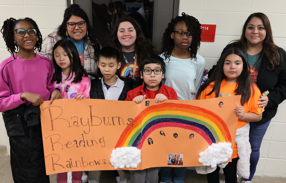 A group of diverse individuals, including children and adults, are holding a sign that reads "Rainbows Reading Rainbows" against a backdrop of a school hallway.