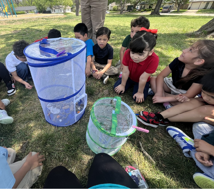 A group of children gathered around a large transparent container, engaged in an outdoor activity on a grassy field surrounded by trees.