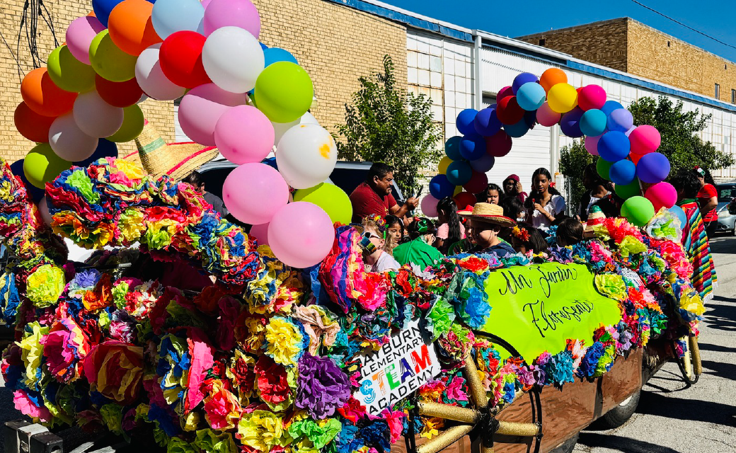 A vibrant and colorful street scene with a crowd of people surrounded by an array of balloons, flowers, and festive decorations, set against a backdrop of brick buildings.
