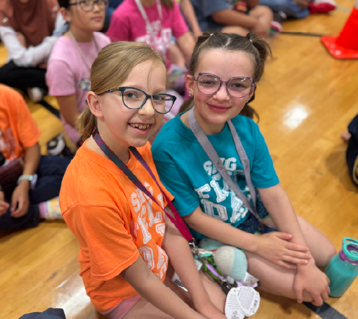 Two young girls wearing colorful shirts and glasses are sitting together and smiling in what appears to be a classroom or educational setting.