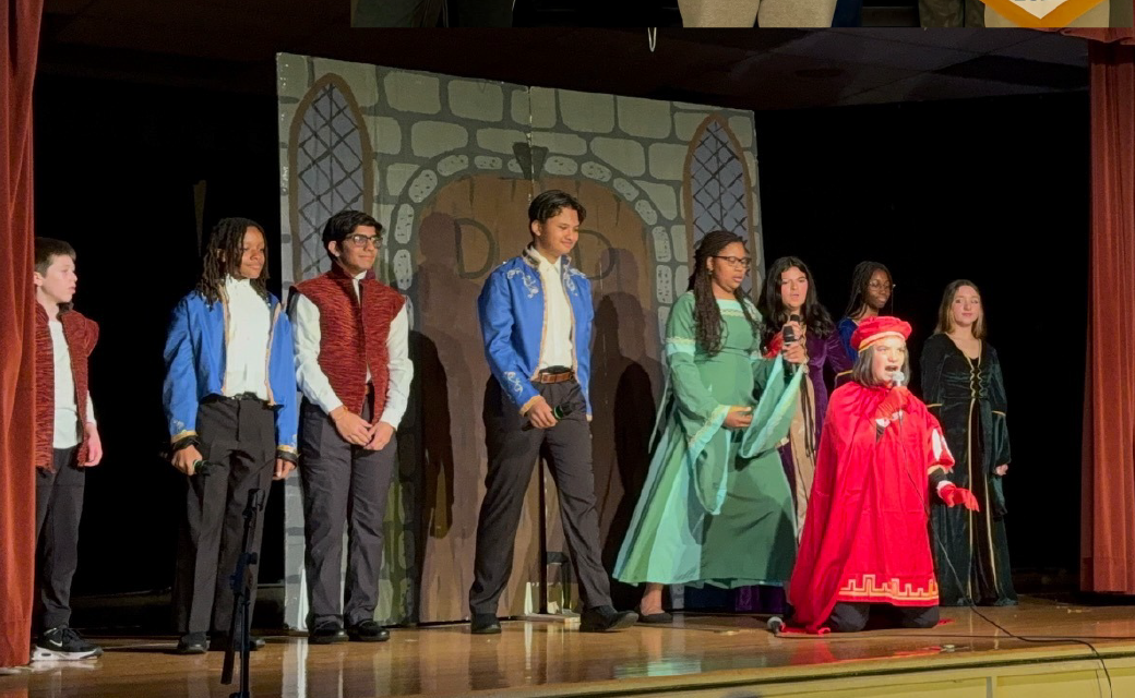 A group of people in various colorful costumes and outfits standing on a stage with a stone archway backdrop.