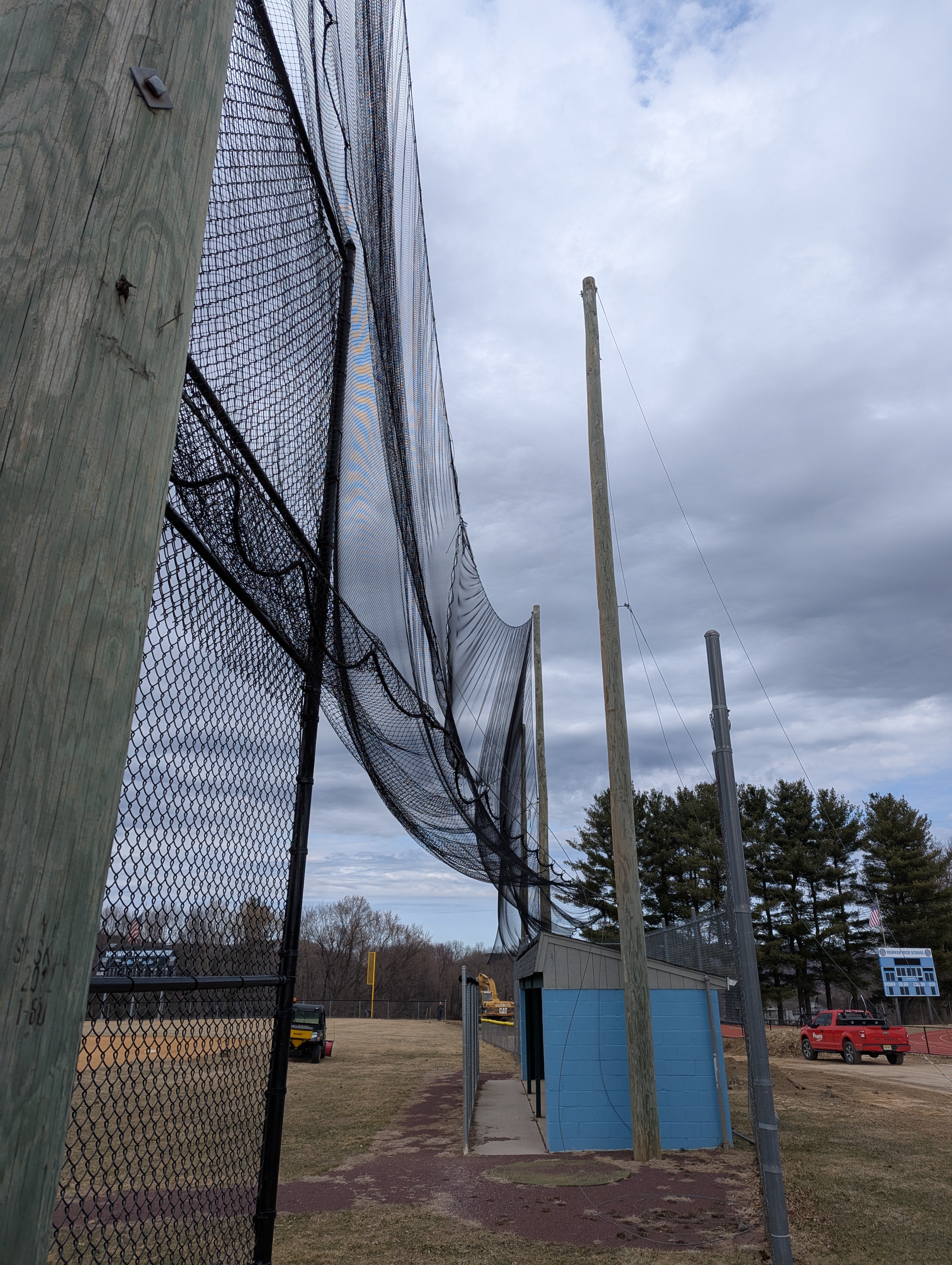 A black safety net stretches across a baseball field with a small blue dugout in the background under a cloudy sky.