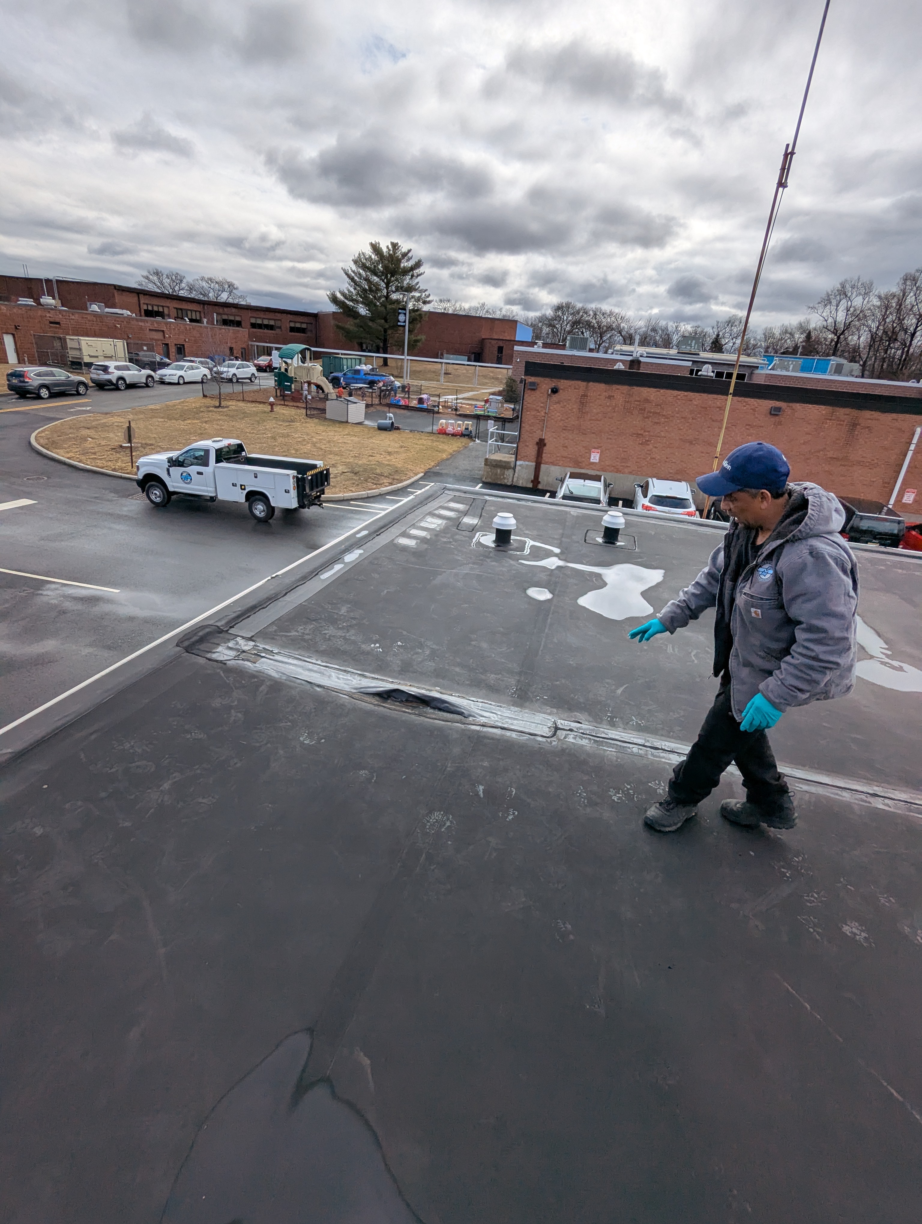 A worker in the foreground points to a damaged roof section, while in the background, a white pickup truck is parked near a brick building under a cloudy sky.